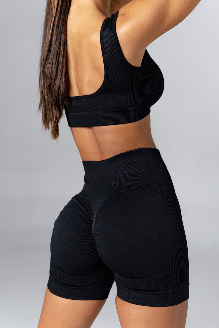 Mujer usando conjunto deportivo seamless negro push up con top y short, con mancuernas rosas y botella de agua negra.