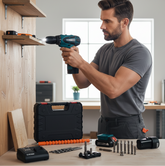 Un hombre barbudo, de complexión fuerte y vestido con una camiseta gris, está utilizando el taladro atornillador inalámbrico de color turquesa y negro para fijar un estante de madera clara a la pared. La escena se desarrolla en un taller bien iluminado con una ventana de fondo. En primer plano, sobre la mesa de trabajo, se encuentran la caja de transporte negra del taladro, una batería adicional, el cargador, un surtido de puntas y brocas, y un extensor de eje naranja.